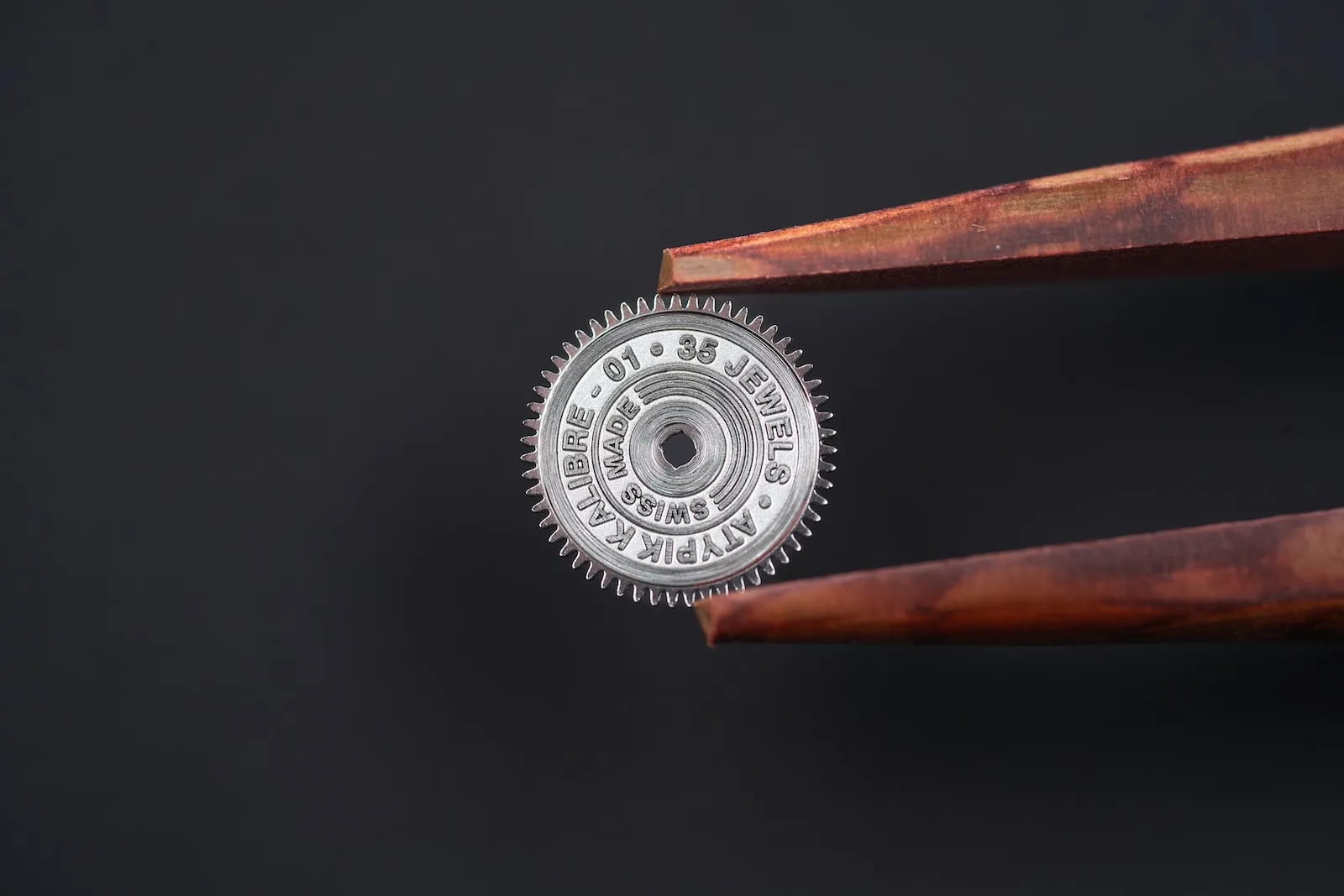 Laser-engraved Atypik Kalibre 01 ratchet wheel held in tweezers